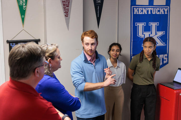 photo of UK students presenting A small group stands in a campus room as a student presenter gestures while explaining a project, with University of Kentucky banners and displays visible in the background.