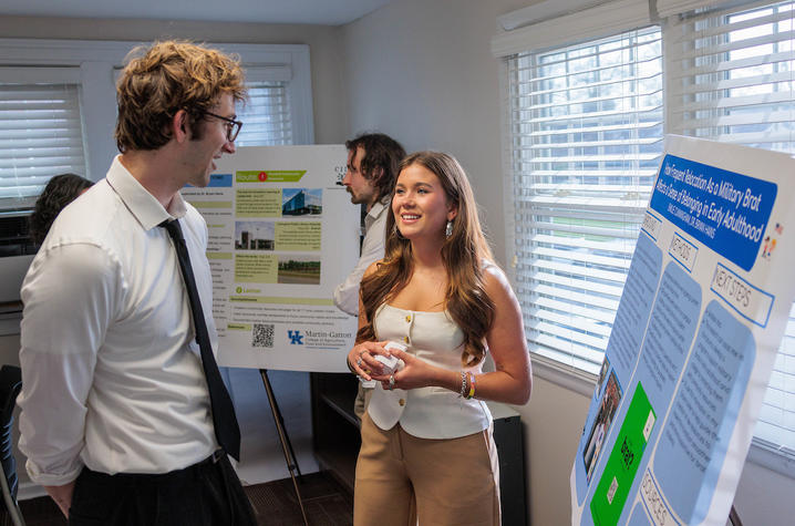 photo of UK student presenting Two people discuss a research poster during a poster session, with additional presentation boards displayed in the room behind them.