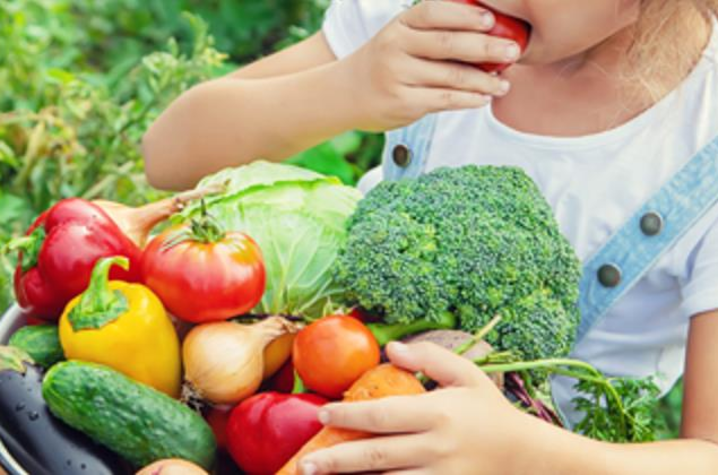 A child holds a large bowl filled with fresh vegetables — including tomatoes, broccoli, bell peppers, cucumbers, onions, and lettuce — while taking a bite of a tomato in a garden setting.