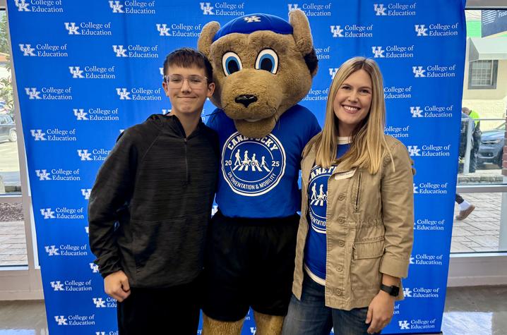 University of Kentucky backdrop with two people posing beside a costumed mascot wearing a blue shirt and cap featuring a circular logo.