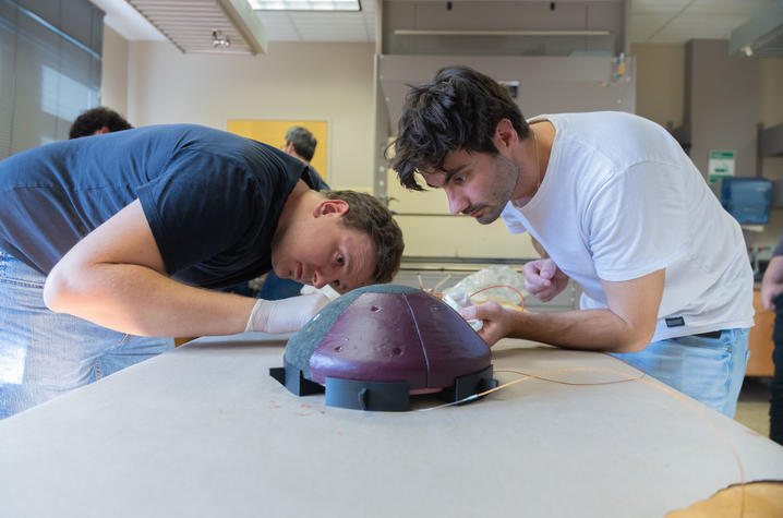 Two students lean over a table working closely on a small dome-shaped device in a classroom or lab setting.