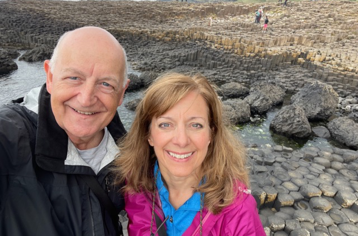 A couple takes a selfie while standing among the interlocking basalt columns at a rocky coastal landscape. The ocean is visible in the background, along with cliffs and scattered visitors exploring the area.