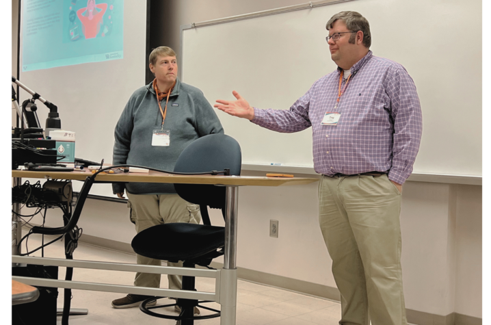 Christopher Huggins and Troy Cooper in front of a whiteboard doing a presentation. A projector screen is visible to the side. 