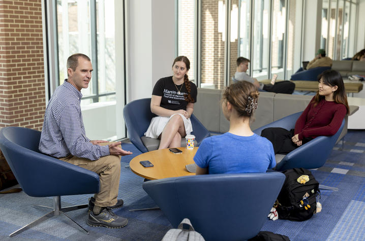Four people sit in blue lounge chairs around a small round table inside a bright, modern building with large windows, brick walls, and other seated people visible in the background.