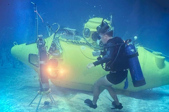 Photo of Nekton Gamma submersible A scuba diver wearing diving gear interacts with a yellow underwater submersible on the ocean floor, surrounded by blue water and scientific equipment illuminated by lights.