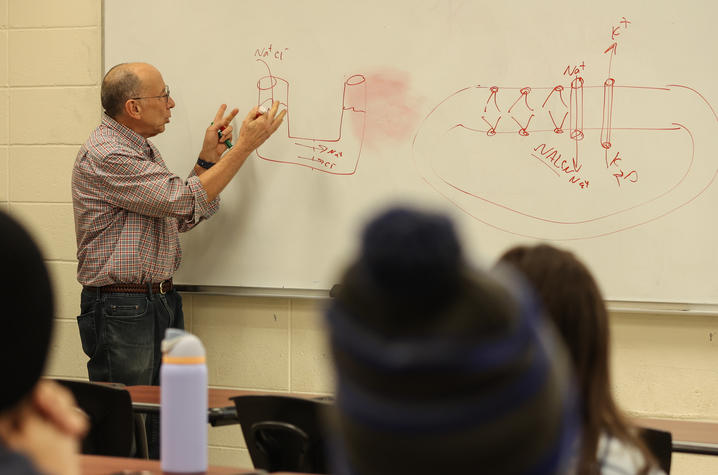 A professor teaches in front of a classroom whiteboard covered with scientific diagrams and notes while students listen from their seats.