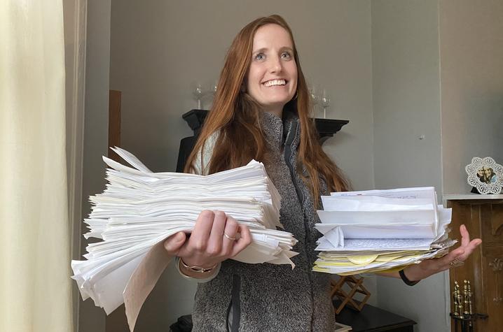 photo of UK alum Rosie Paulik A person standing indoors holds large stacks of white papers and envelopes in both arms, with documents fanned out and slightly overflowing, suggesting a heavy workload or paperwork overload.