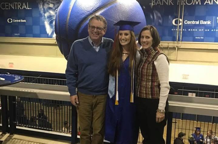 A graduate in a blue cap and gown stands between two adults, all smiling and posing for a photo inside an indoor arena with University of Kentucky banners and a large basketball display in the background.