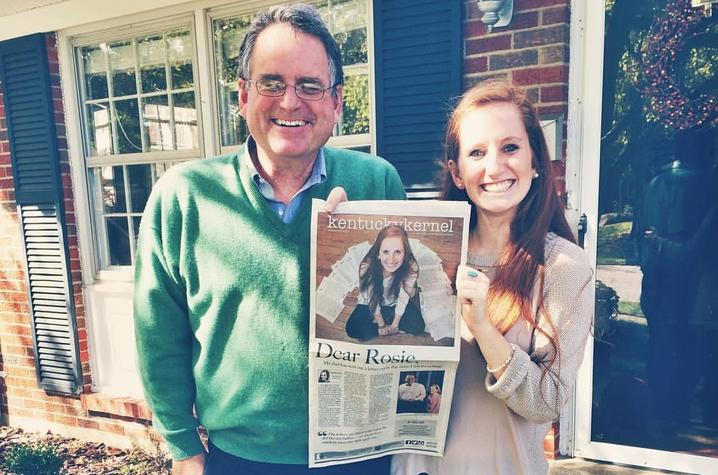 Two people stand outdoors in front of a brick house, smiling and posing together. One person holds up a newspaper featuring a large photo on the front page, while the other stands beside them with an arm around the newspaper holder.