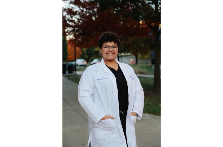 Person wearing a white lab coat stands outdoors on a sidewalk with hands in pockets, smiling, with trees showing fall colors in the background.