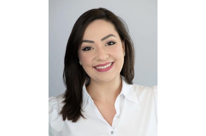 Head-and-shoulders professional portrait of a woman with dark shoulder-length hair, smiling at the camera, wearing a white blouse against a light gray background.