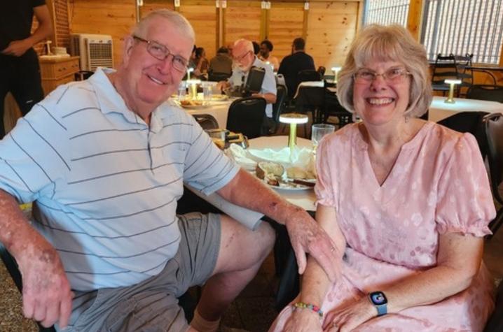 Photo of paitent Kathie Kok with her husband. Two people seated at a restaurant table, posing for a photo in a dining area with other patrons visible in the background.