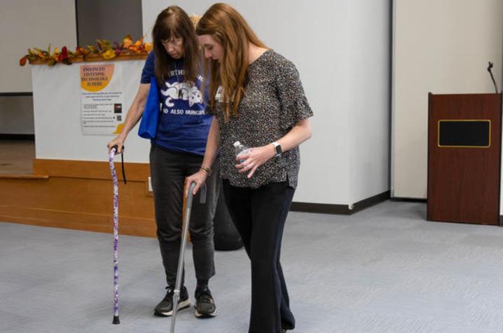 Photo from fall prevention day Person using forearm crutches walks across an indoor room while another person walks beside them, appearing to guide or assist with movement practice.