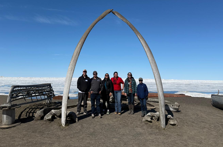 Six people stand beneath a large arch formed by two whale bones on a dark shoreline, with wooden boats and a vast expanse of sea ice beneath a clear blue sky in the background.