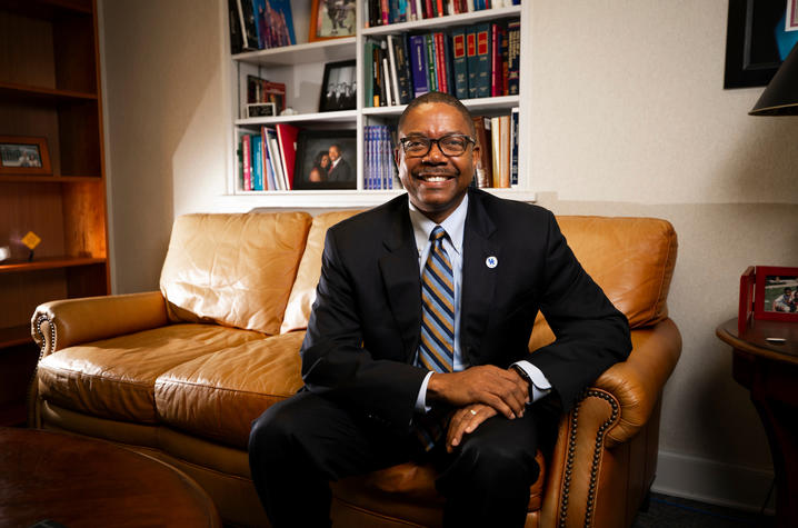 Dean Brennen seated in office on brown couch wearing suit and glasses