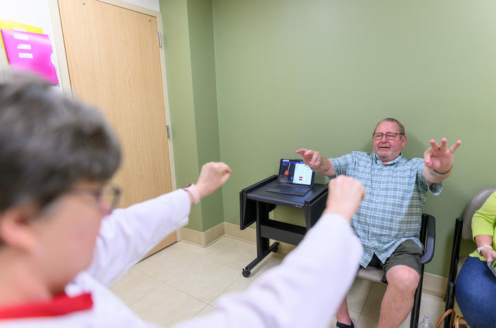 An older male research participant sits in a chair, his arms extended in a small exercise. In front of him, a woman with gray hair wearing a white medical coat stands in front of him with her arms extended, guiding him in the exercise. 