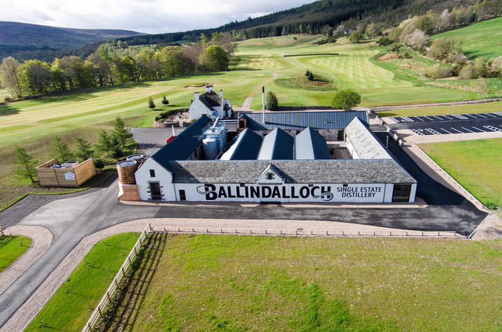 Aerial view of Ballindalloch Single Estate Distillery, showing white stone buildings with slate roofs, surrounding paved drives, and green fields, set amid rolling hills, trees, and patchwork farmland under a bright sky.