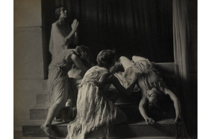 image of Dancers in Company Black-and-white photograph of several figures in flowing garments posed on steps, some bent or reaching while one stands upright with raised hands. The scene resembles a staged performance or expressive dance in low, dramatic lighting.