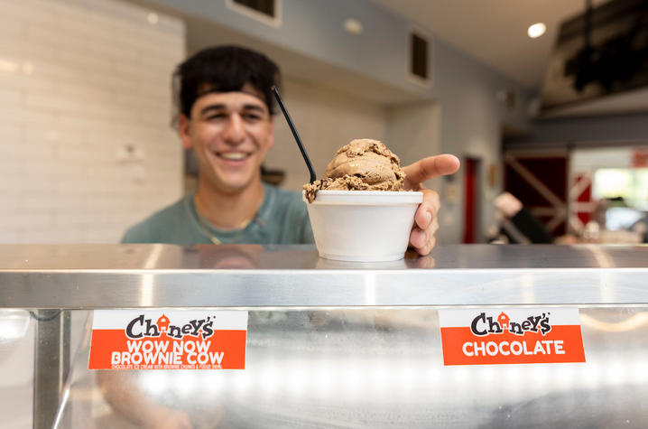 photo of Daniel Carneiro A person stands behind an ice cream display case, holding out a cup filled with two scoops of chocolate ice cream and a spoon. Flavor labels on the case read “Chaney’s Wow Now Brownie Cow” and “Chaney’s Chocolate.”