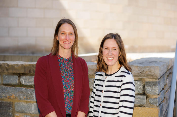 Two people standing next to each other to pose for a picture. One person is wearing a maroon blazer and the other a white and black striped cardigan 