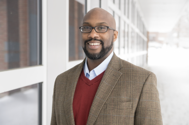 A person stands outdoors near a building with large windows, wearing a brown plaid blazer layered over a red sweater and a collared shirt. Snow covers the ground in the background.