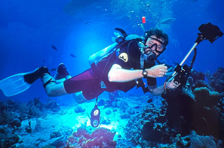 Photo of Greg Gerhardt during a dive A scuba diver swims above a coral reef in clear blue water while holding an underwater camera rig.