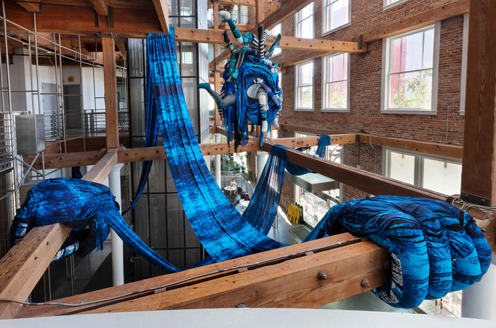 Large blue fabric sculpture draped across wooden beams in a gallery space.