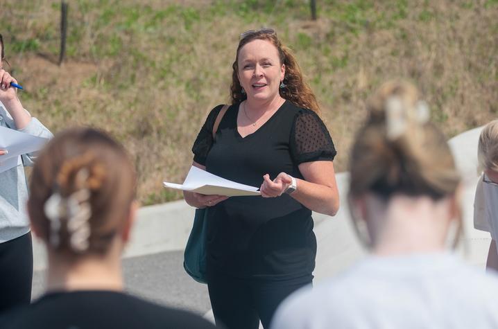A person stands outdoors holding papers and speaking to a small group, with several people listening in the foreground and a grassy hillside in the background.