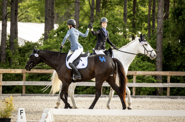 Eventing Team shaking hands Two riders in helmets and formal equestrian attire ride side by side in an outdoor dressage arena, reaching across to clasp hands while mounted on a dark horse and a white horse. The horses wear competition number tags.