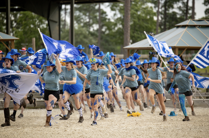Cheering at a competition A large group of University of Kentucky supporters runs across an outdoor equestrian venue carrying blue and white UK flags and banners. Participants wear matching blue hats and light blue shirts, with blue body paint and accessories, as they move.