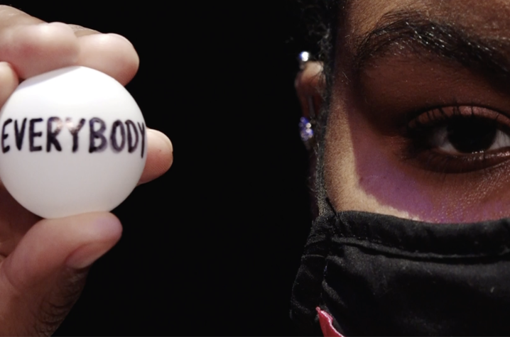 a close-up photo of masked student's fingers holding a ping pong ball reading EVERYBODY beside her eye