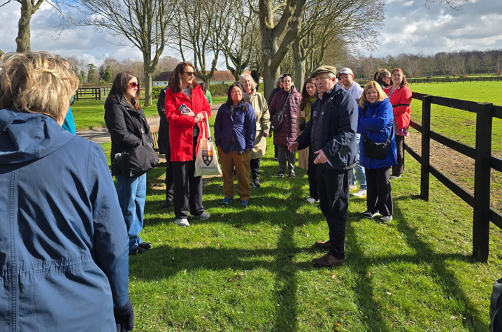 A medium group of people standing around listening to a person speaking on green pasture with a fence in the background.