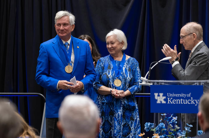 Two people on a stage at the University of Kentucky hold medallions while another person applauds at a podium with a UK logo.