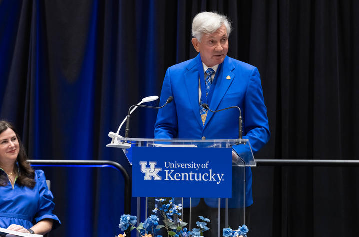 A person in a blue jacket speaks at a University of Kentucky podium on a stage with blue-and-white floral arrangements.