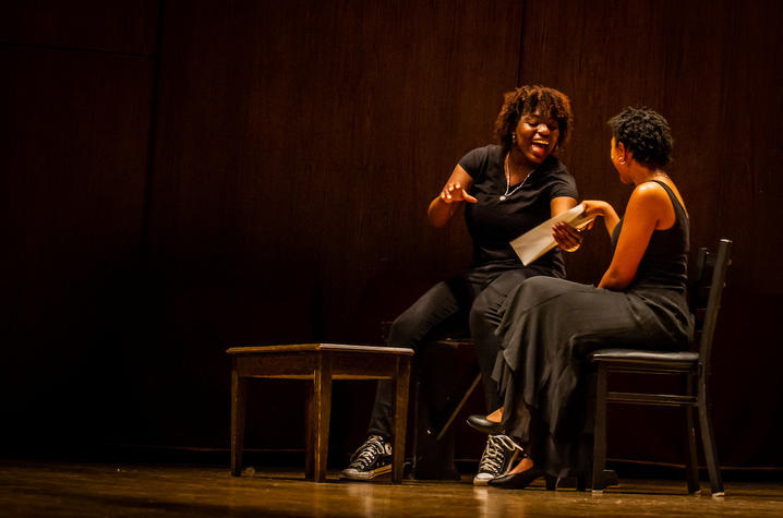 Two black women seated next to each other on state, performing a dramatic play.