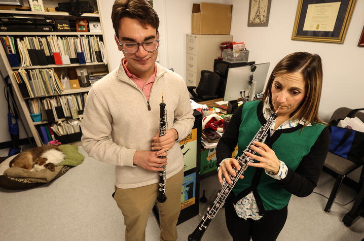 Two people hold oboes in a music office. One person is playing and another is watching.