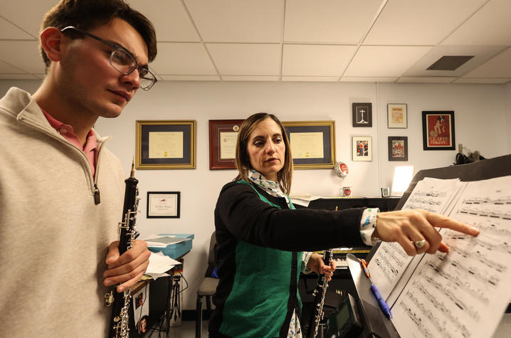 Person points to sheet music while standing beside another person holding an oboe in a music office.