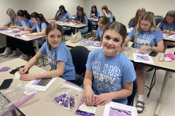 Students sit at desks placing purple and black LEGO bricks to help build a butterfly art display.