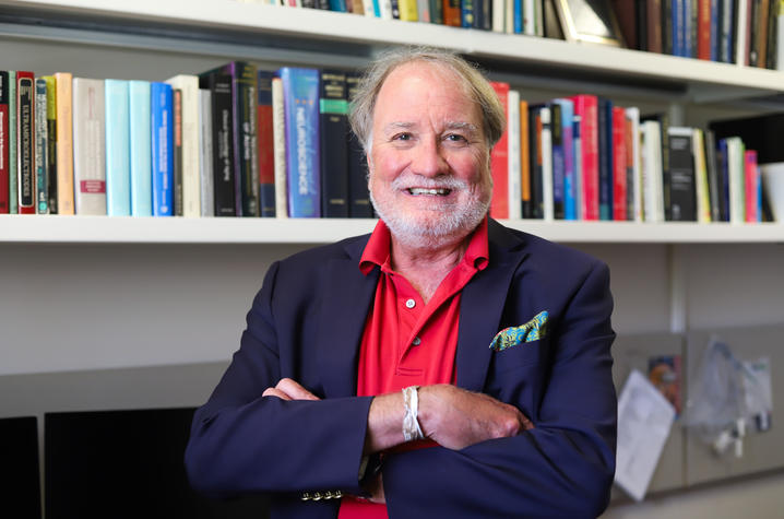 Photo of Greg Gerhardt A person wearing a navy blazer over a red shirt stands with arms crossed in a book-filled office, with shelves of colorful books and papers visible in the background.