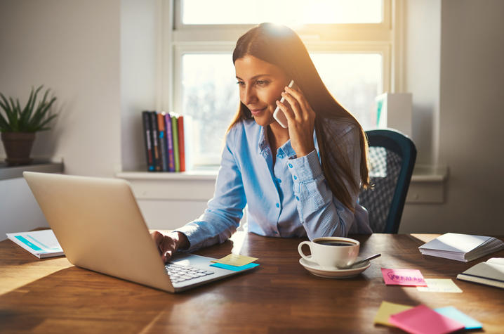 photo of woman sitting at table at home with laptop and cell phone