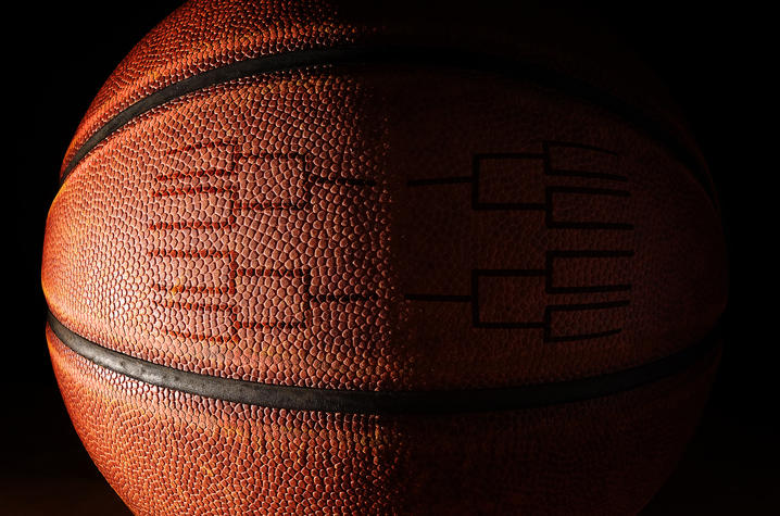 A close‑up photograph of a basketball set against a dark background. The ball is lit from the left side, revealing its textured surface.