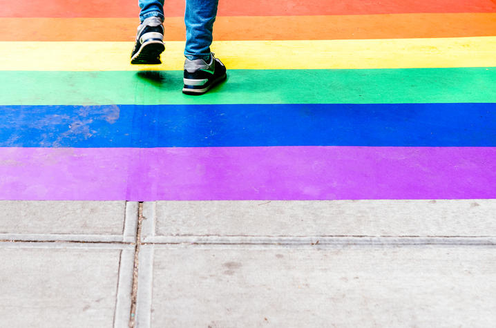 The image shows the lower legs of a teenager walking over a rainbow sidewalk; they're wearing jeans and black sneakers.