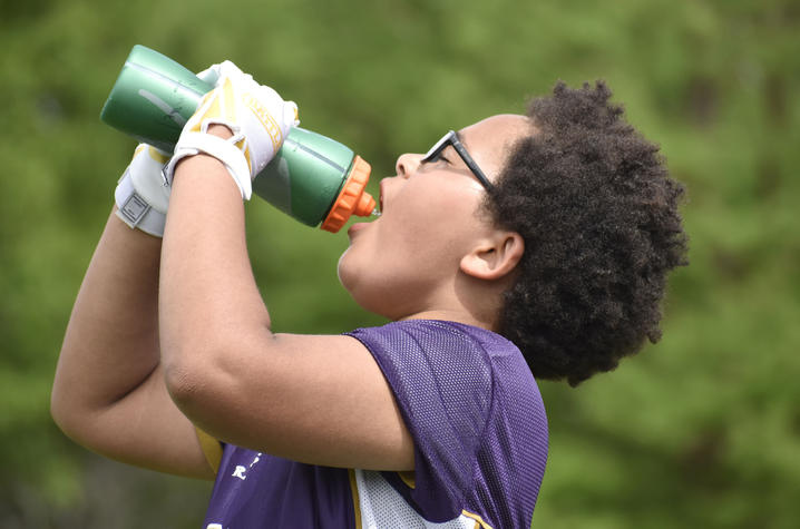 Student athlete hydrates during outdoor activity. Student athlete wearing a football practice jersey and gloves drinks water from a sports bottle outdoors during warm weather.