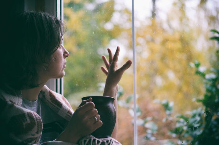 A person holds a mug while looking out a window at autumn foliage, with one hand raised toward the glass.