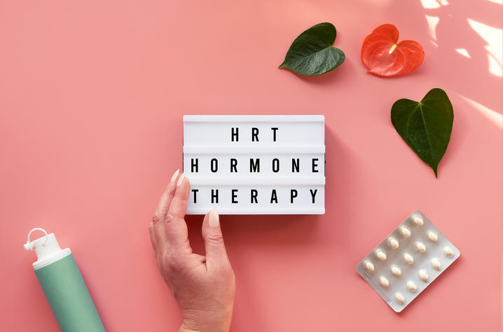 A hand holds a lightbox sign reading “HRT Hormone Therapy” surrounded by tropical leaves, pills, and a pump bottle on a pink background.