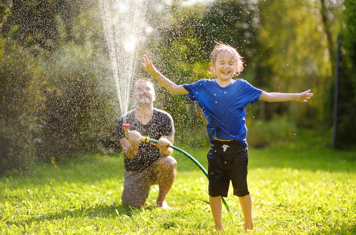 Stock photo of child running through sprinkler spray while an adult cools the yard with a garden hose. An adult kneels in a grassy yard holding a garden hose while spraying water overhead as a child in a blue shirt and dark shorts runs through the spray with arms outstretched on a sunny summer day.