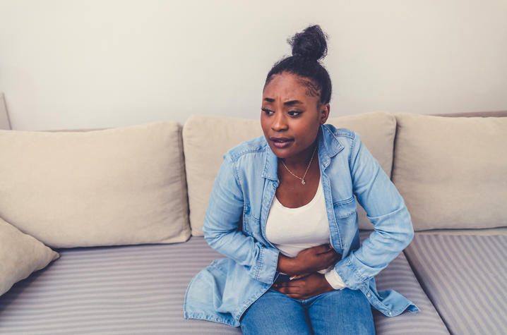 Person sitting on a couch indoors, leaning forward and holding their lower abdomen with both hands.