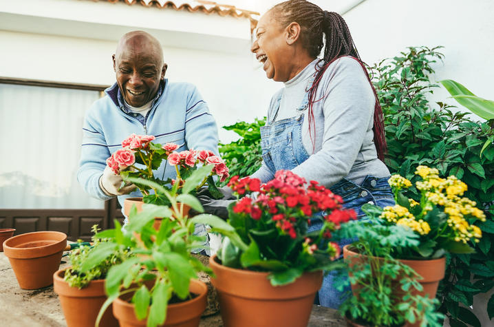Two older adults sit at an outdoor table surrounded by terracotta pots of colorful flowers and plants, smiling and talking while tending to a small container garden.