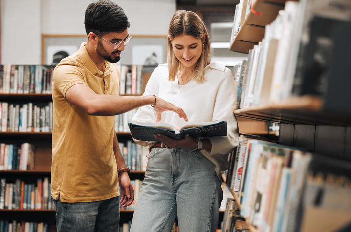 Two people looking at a book at a bookshelf. 