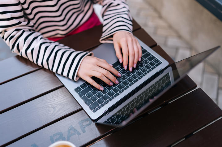 Person wearing a striped long‑sleeve shirt typing on a laptop at an outdoor wooden table.